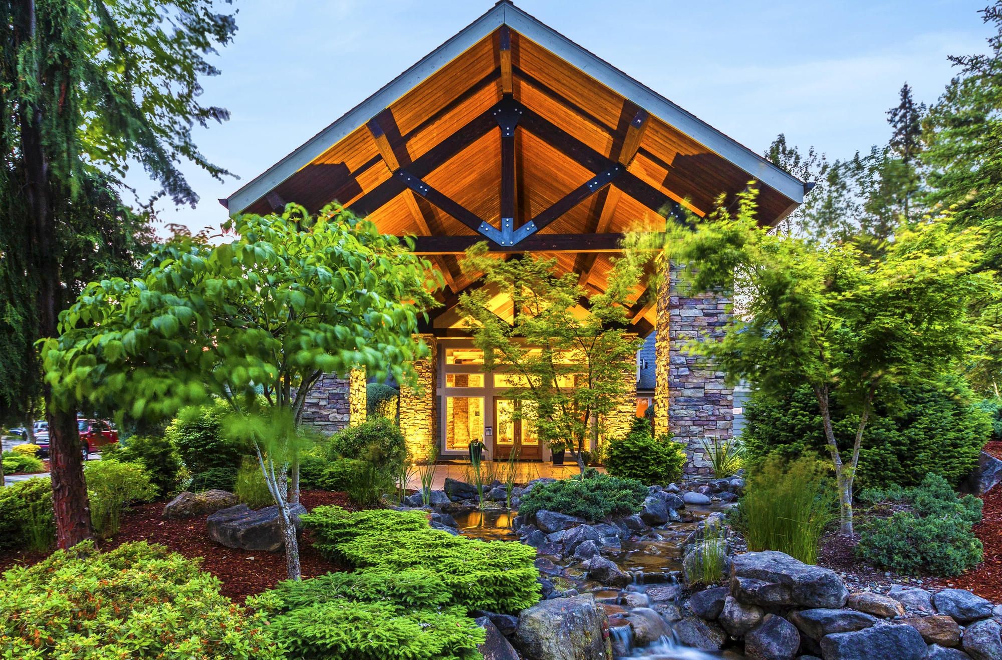 Modern wooden house with large front windows, surrounded by lush greenery and a landscaped garden with rocks.