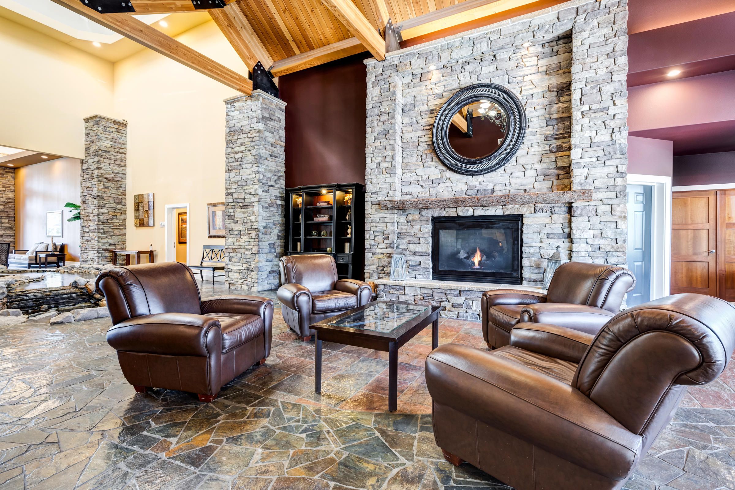 Spacious living room with stone fireplace, leather chairs, glass table, and vaulted wood ceiling.