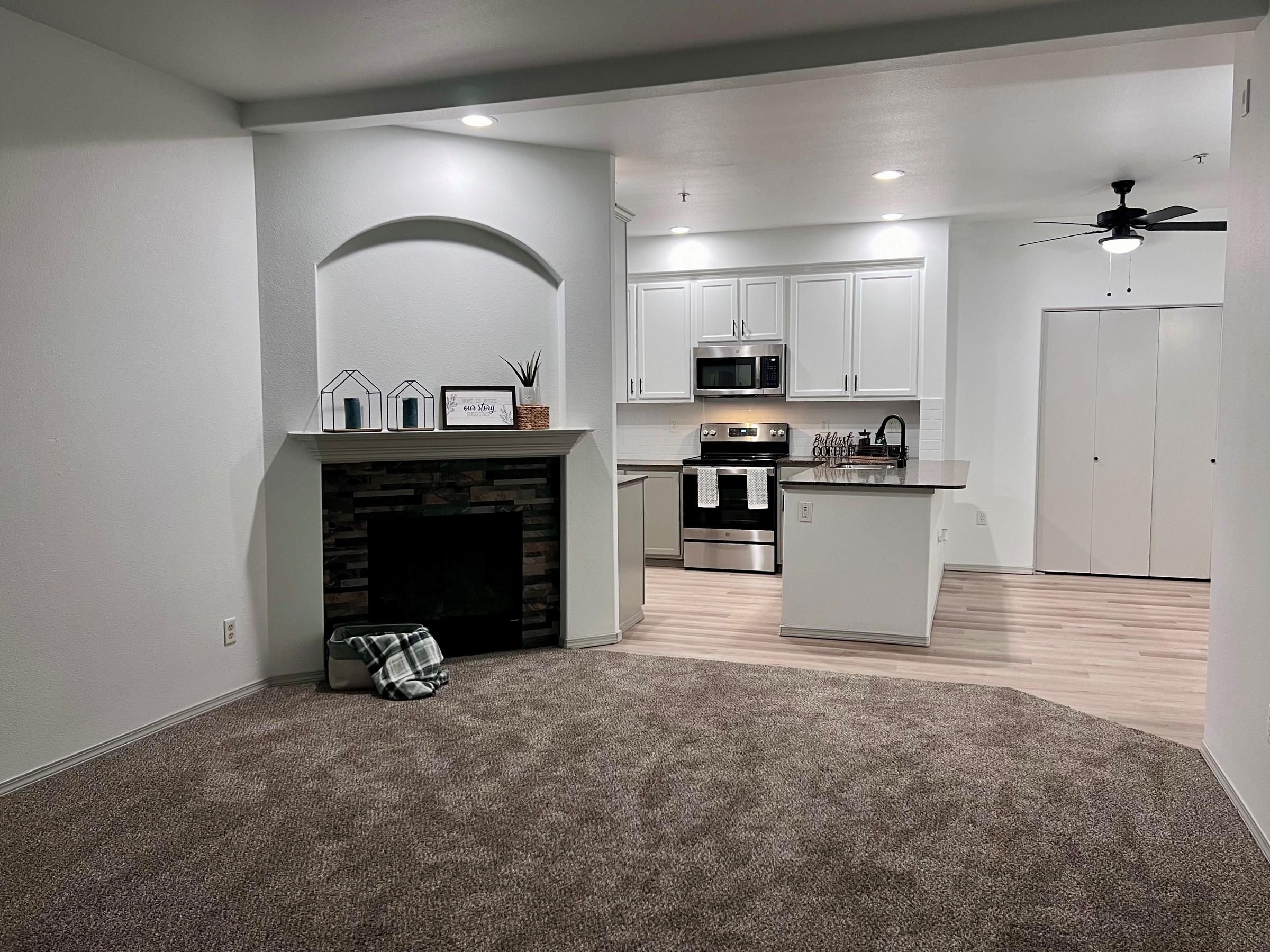Modern living room with carpet, a fireplace, and an open kitchen with white cabinets and stainless steel appliances.