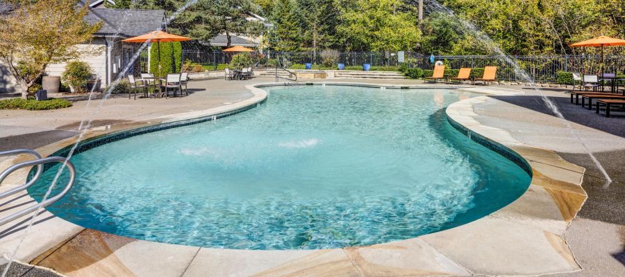 Outdoor swimming pool with water fountains, lounge chairs, and orange umbrellas surrounded by trees.