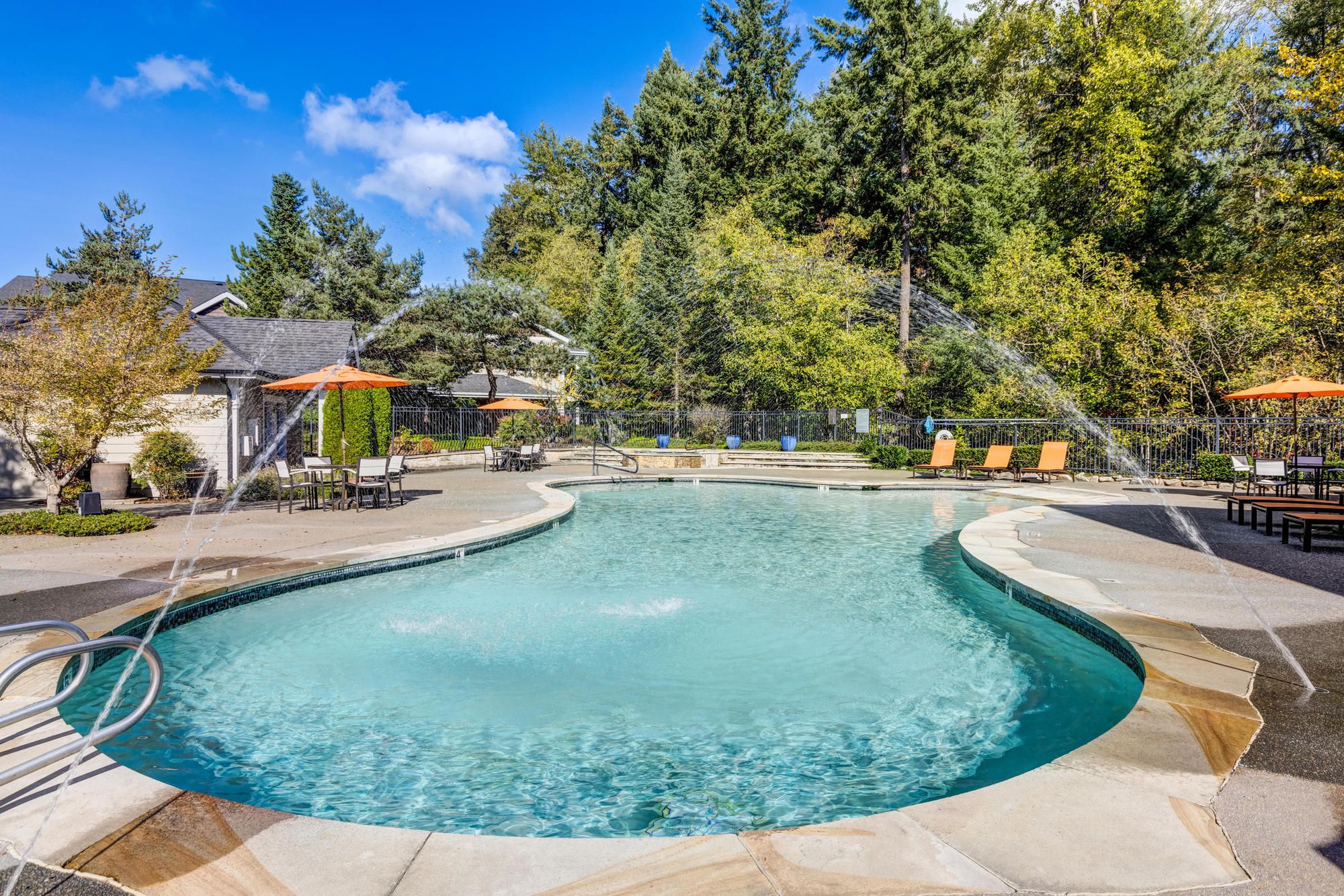 Outdoor swimming pool with water fountains, lounge chairs, and orange umbrellas surrounded by trees.