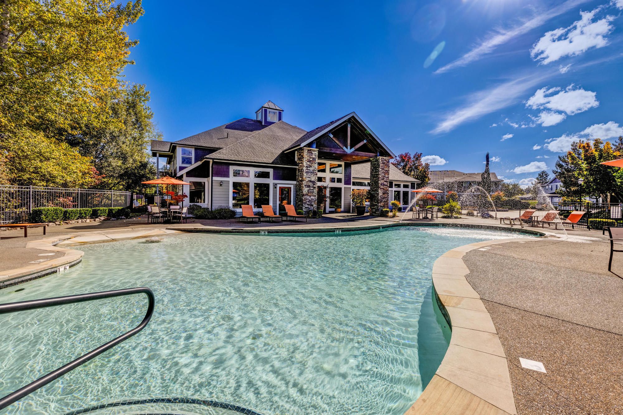 Outdoor pool with lounge chairs beside a modern clubhouse under a clear blue sky with scattered clouds.