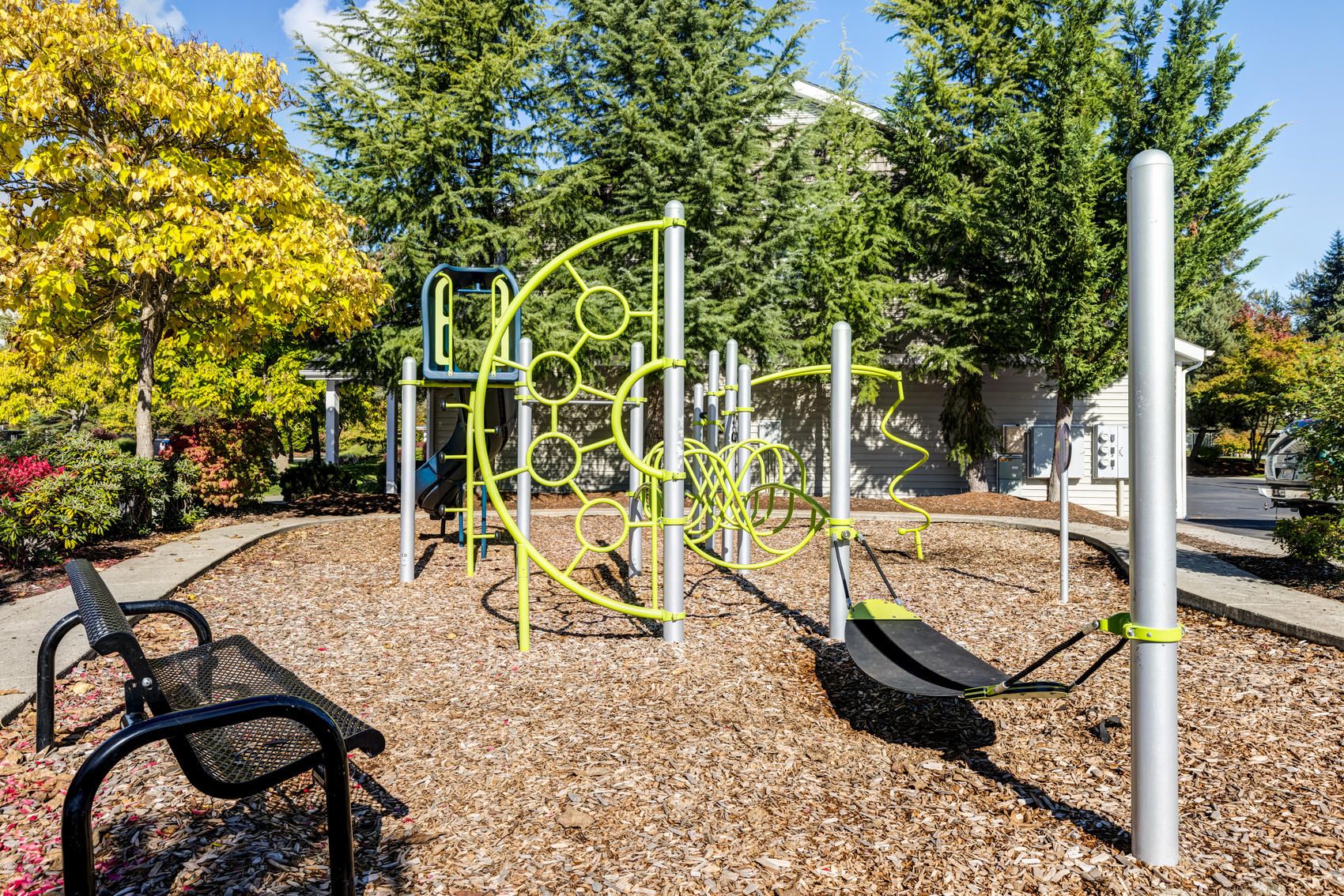 Modern playground with climbing structures and benches, surrounded by trees and wood chips on a sunny day.